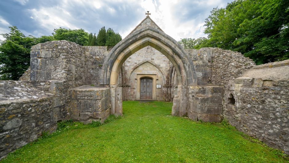 Photograph of an ancient stone church entrance surrounded by grassy area and green trees, with large stone walls on either side. The pointed arch doorway features detailed stone carvings, and a wooden door is visible within the arch. The scene appears to be part of a historic ruin or outdoor heritage site, with natural light illuminating the weathered stonework. This setting offers a tranquil view of medieval architecture, possibly used for educational or tourism purposes, within a natural landscape. The image showcases the structural remains of the church's exterior, emphasizing the stone construction and historical significance, set against a backdrop of leafy trees and a partly cloudy sky. Such historical ruins may be suggestive of locations that require careful handling during house removals or estate cleanouts when in proximity to heritage sites, and the scene aligns with the idea of loading or transport of items in a safe manner for preservation or relocation by professionals like Movers Sutton.
