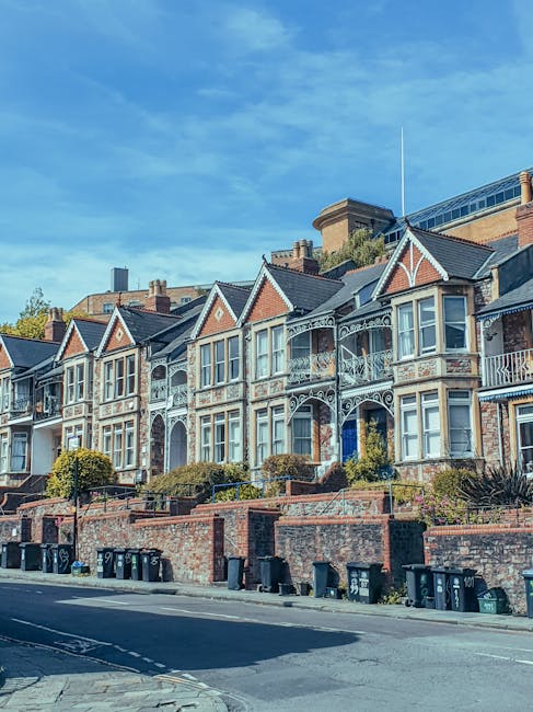 Image of a row of Victorian-style terraced houses situated along a quiet residential street near Sutton station. The houses feature decorative white ironwork balconies, bay windows, and pitched roofs with gable ends. The front gardens are raised and enclosed with brick walls, with some shrubs and small trees visible. On the pavement in front of the houses, several blue and black wheelie bins are lined up. The clear sky above is blue with some wispy clouds. The scene reflects a typical suburban neighbourhood, which might be involved in home relocation or furniture transport, supported by the professional removals services of Movers Sutton, as indicated by the PAGE_TITLE, with the setting ideal for moving and packing activities.